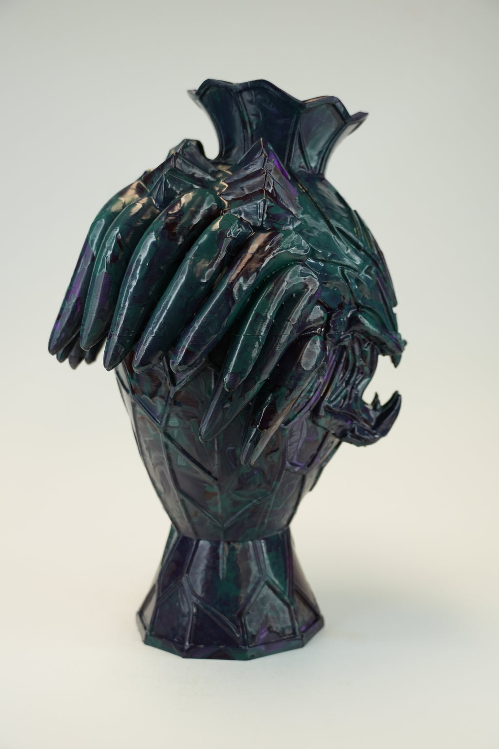 A sculptural ceramic vase with a stylized animal head motif, featuring a dark iridescent glaze with purple, green, and blue tones, set against a plain light gray background.