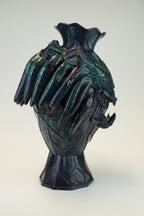 A sculptural ceramic vase with a stylized animal head motif, featuring a dark iridescent glaze with purple, green, and blue tones, set against a plain light gray background.