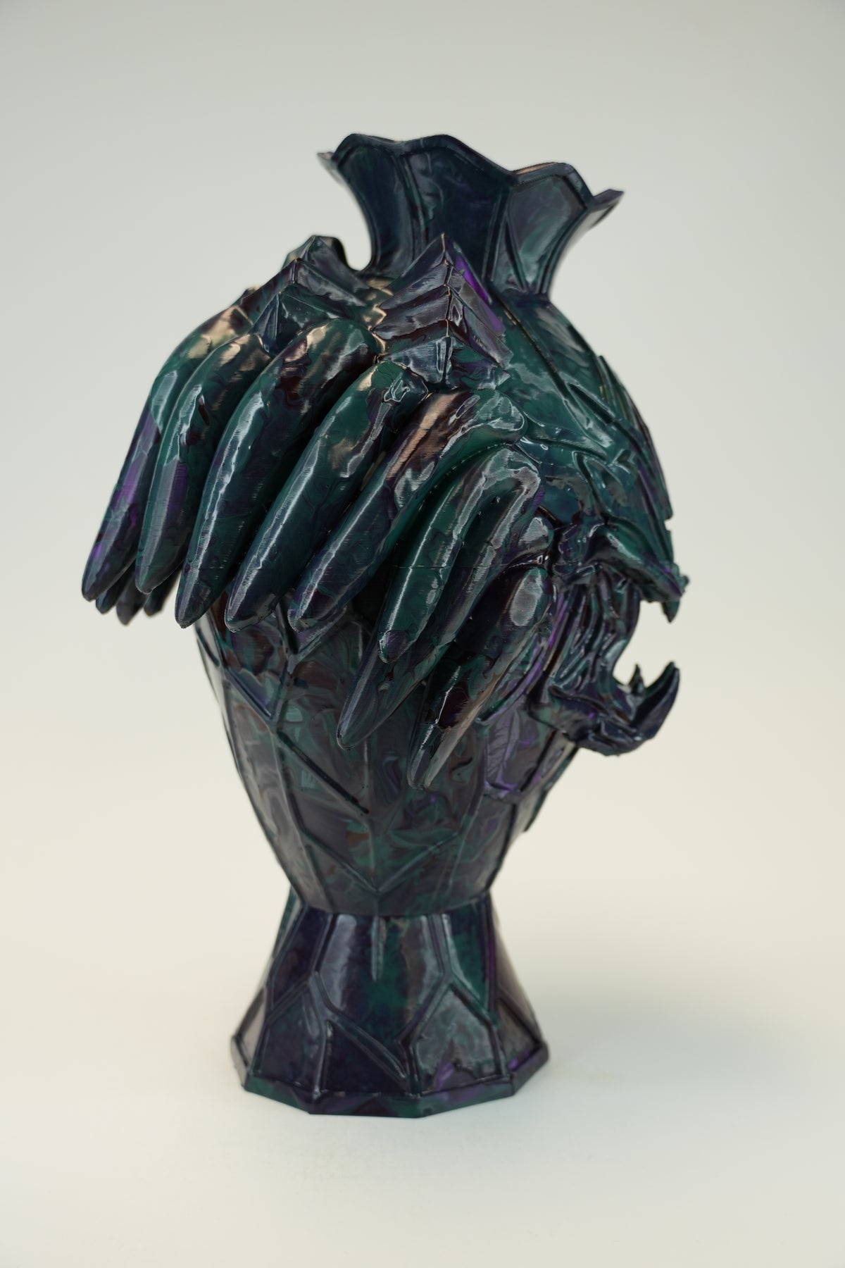 A sculptural ceramic vase with a stylized animal head motif, featuring a dark iridescent glaze with purple, green, and blue tones, set against a plain light gray background.