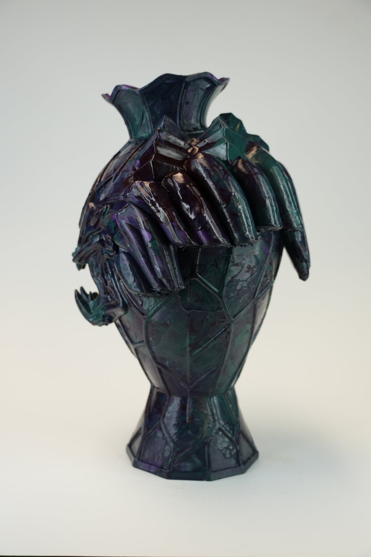 An abstract sculptural ceramic vase with a faceted, geometric design and a flared rim, featuring a dark iridescent finish that shifts between deep purple, blue, and green, set against a plain light gray background.
