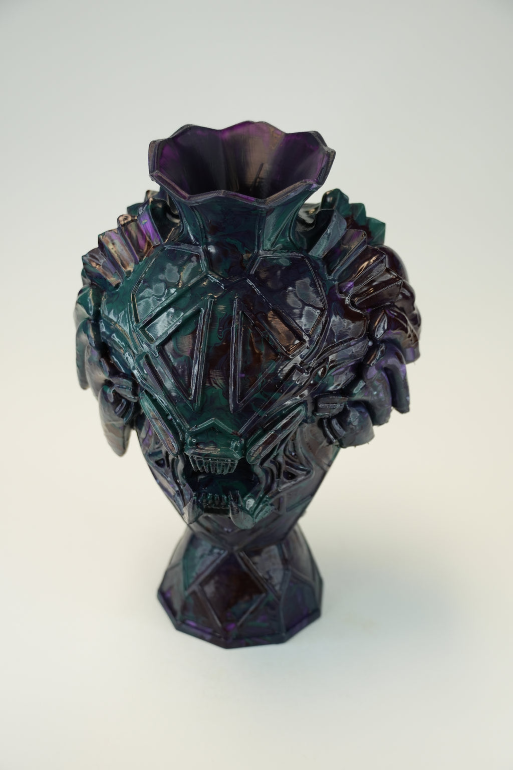 An iridescent glass vase with a sculptural, faceted skull-like design and a flared top, shown on a plain light background.