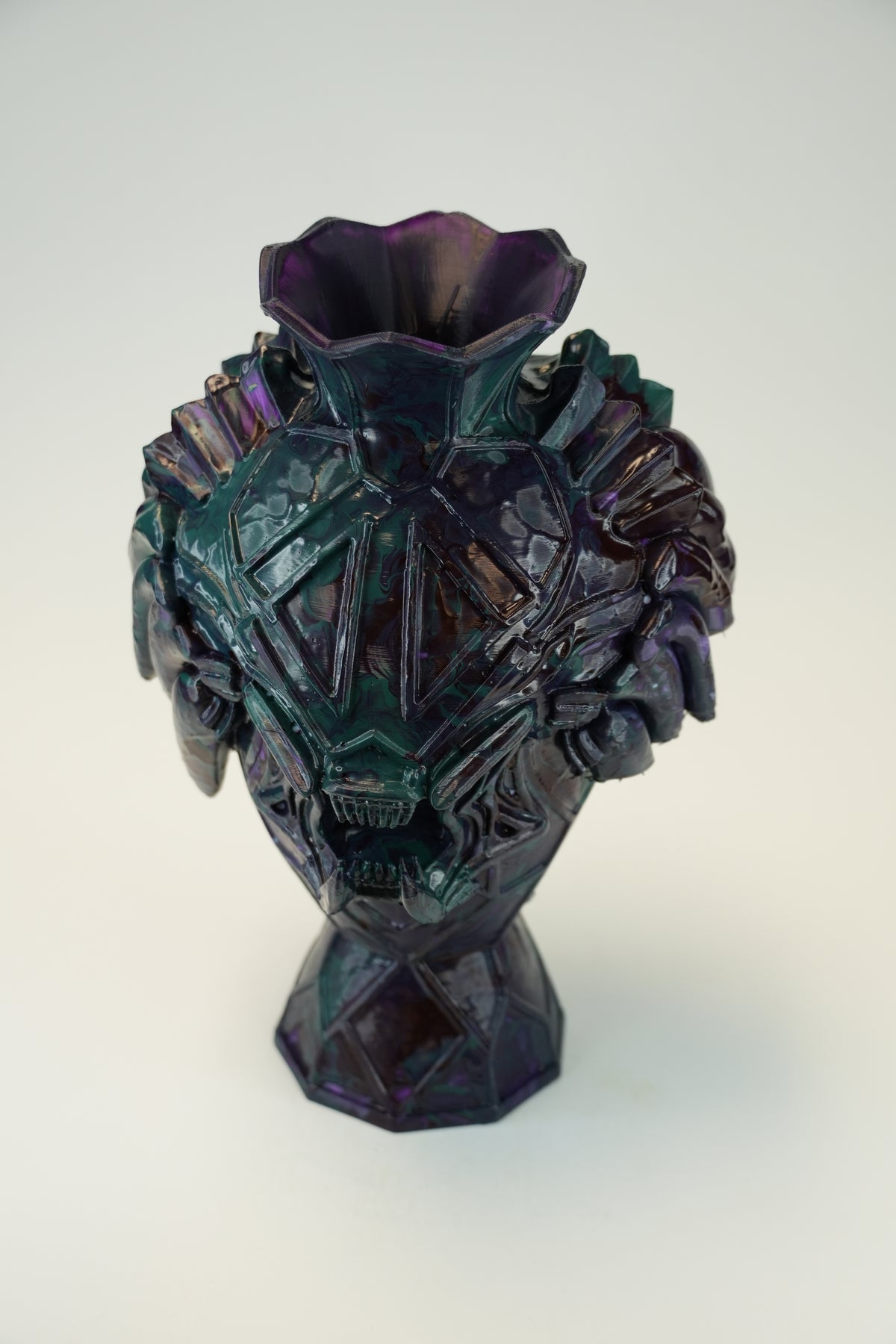 An iridescent glass vase with a sculptural, faceted skull-like design and a flared top, shown on a plain light background.