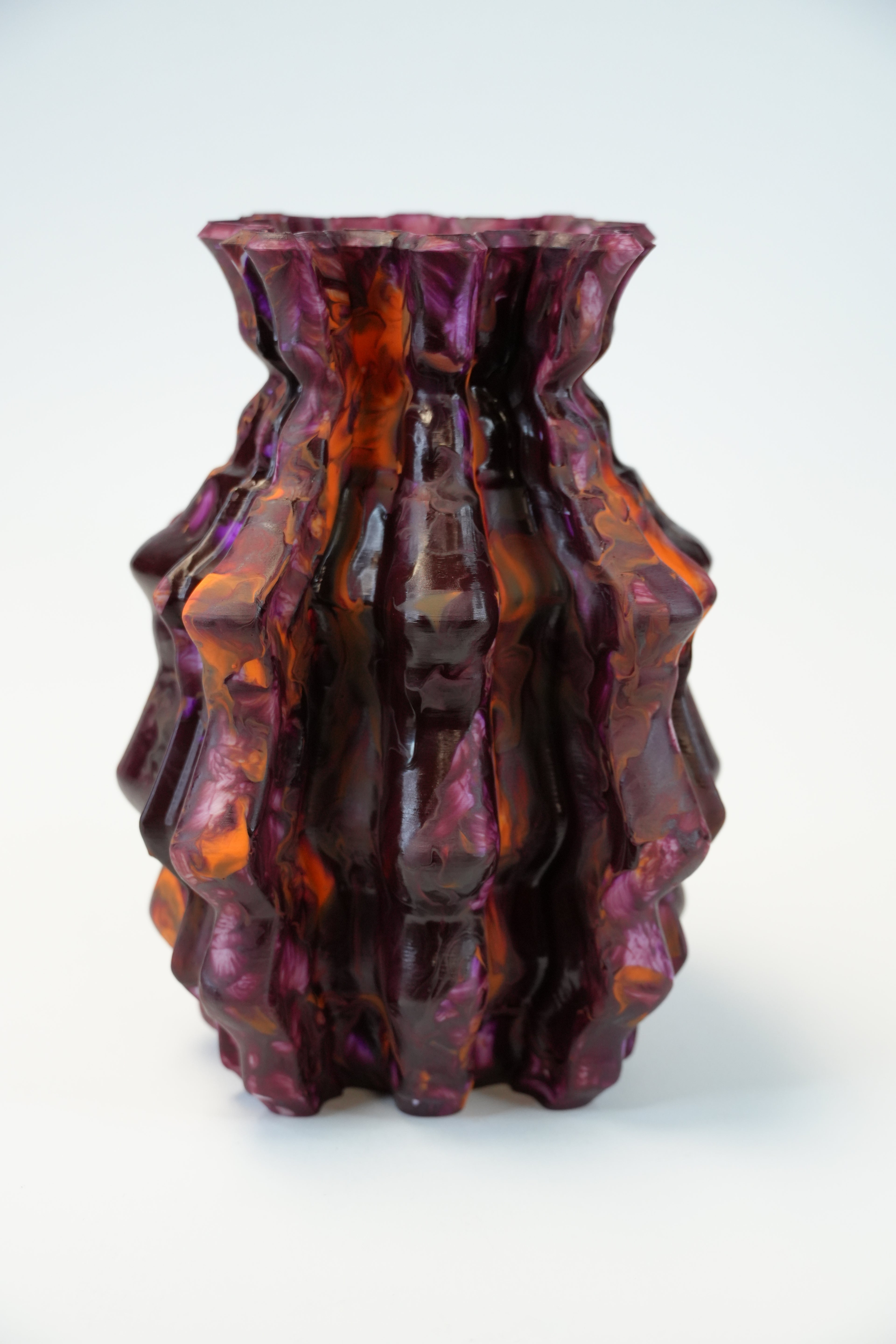 A sculptural, ribbed glass vase with a wavy, organic texture in deep purple and amber-orange tones, set against a plain white background.