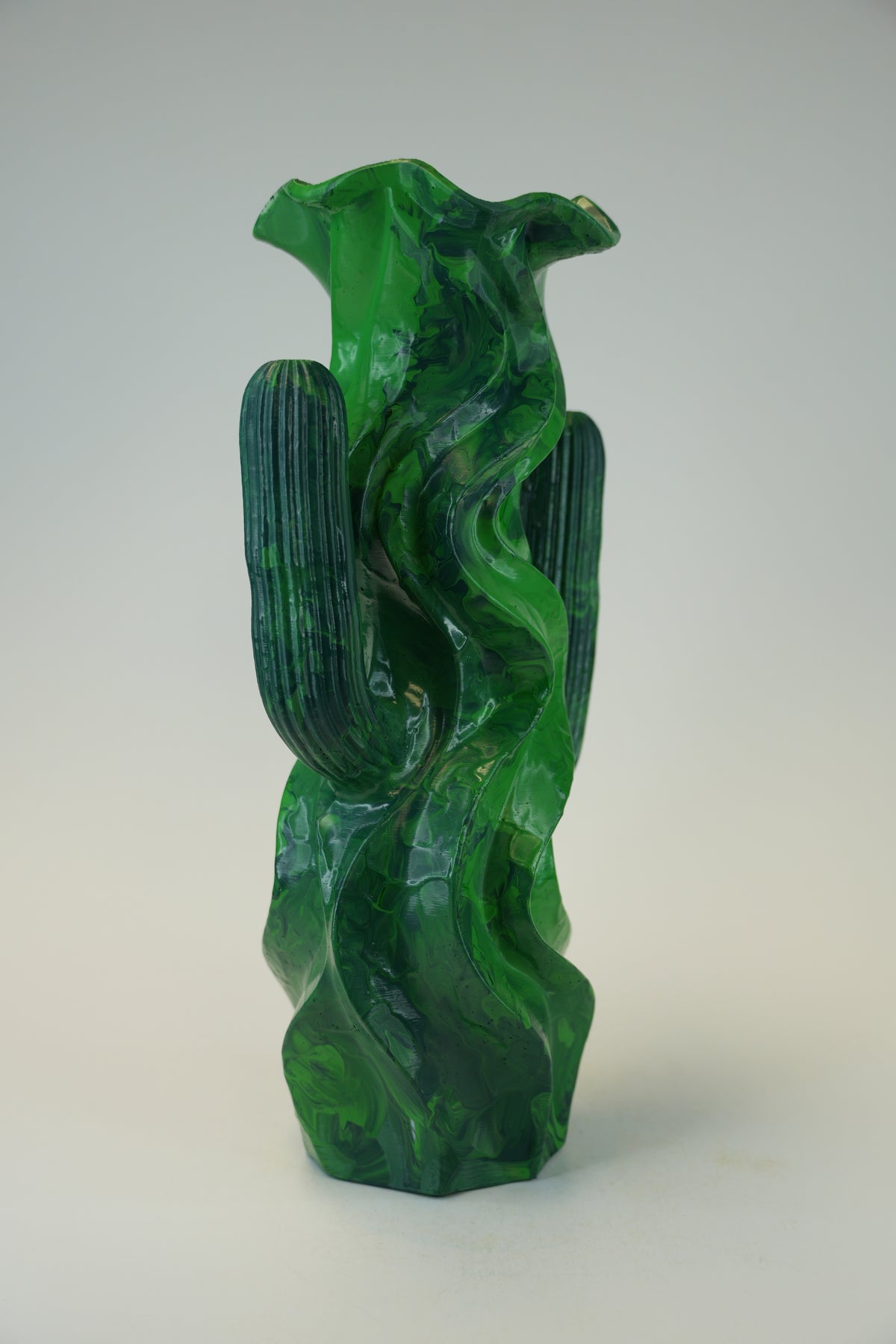 A sculptural green glass vase with a cactus and flowing wave motif, photographed against a neutral light gray background.