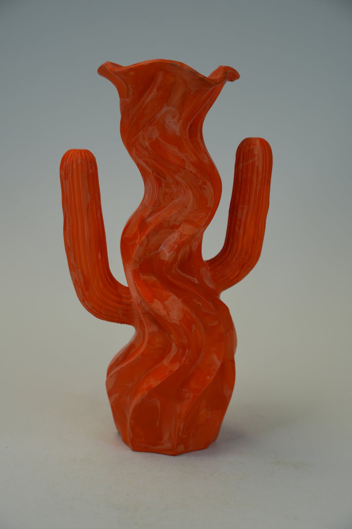 A sculptural ceramic vase shaped like a stylized saguaro cactus, featuring a glossy, ribbed texture in a vibrant terracotta orange-red color, set against a neutral gray background.