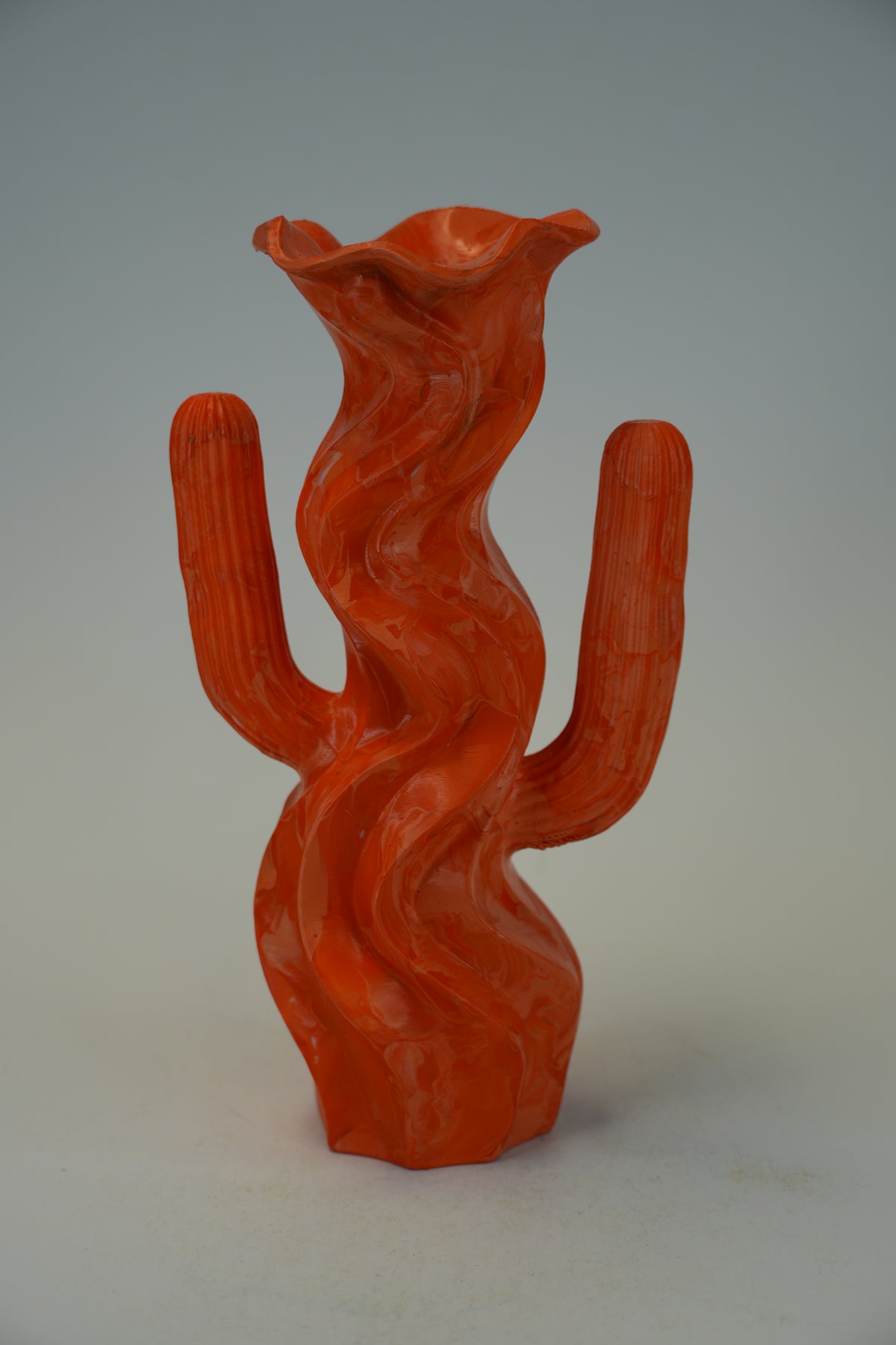 An orange-red sculptural ceramic vase shaped like a stylized cactus with two arms, photographed against a neutral light gray studio background.