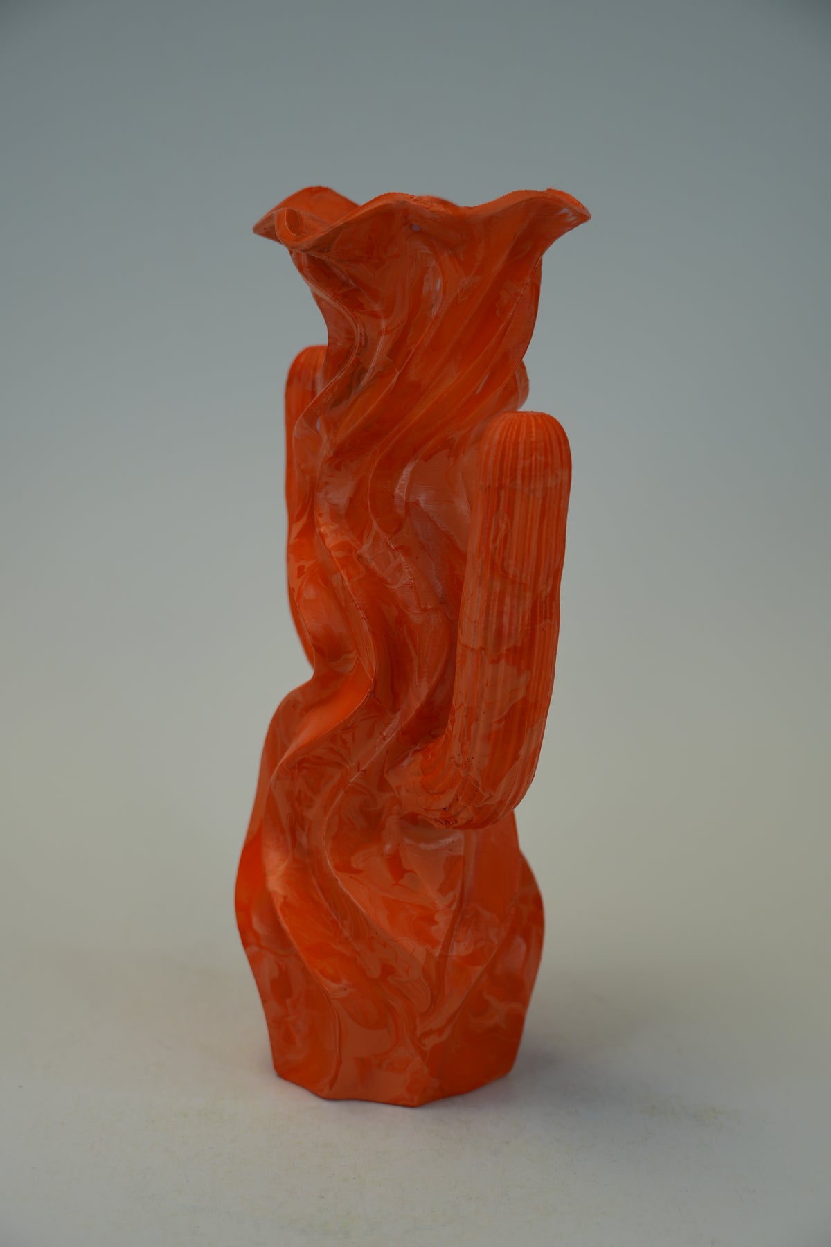 A sculptural orange-red vase with a flared rim and organic, flowing carved lines, photographed against a plain light gray studio background.