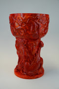 A glossy red sculptural goblet or cup with an ornate carved base featuring a seated figure, photographed against a neutral light gray studio background.