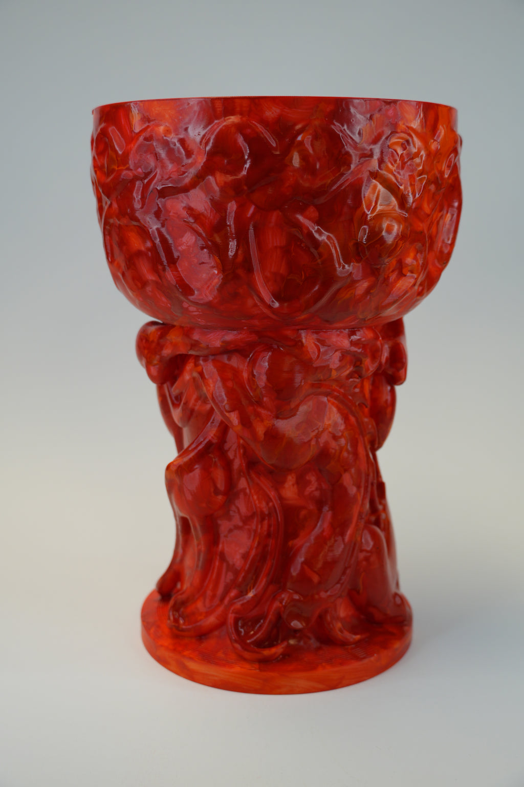 A glossy red glass goblet or chalice with an ornate sculpted base featuring draped figures, photographed against a neutral light gray background.