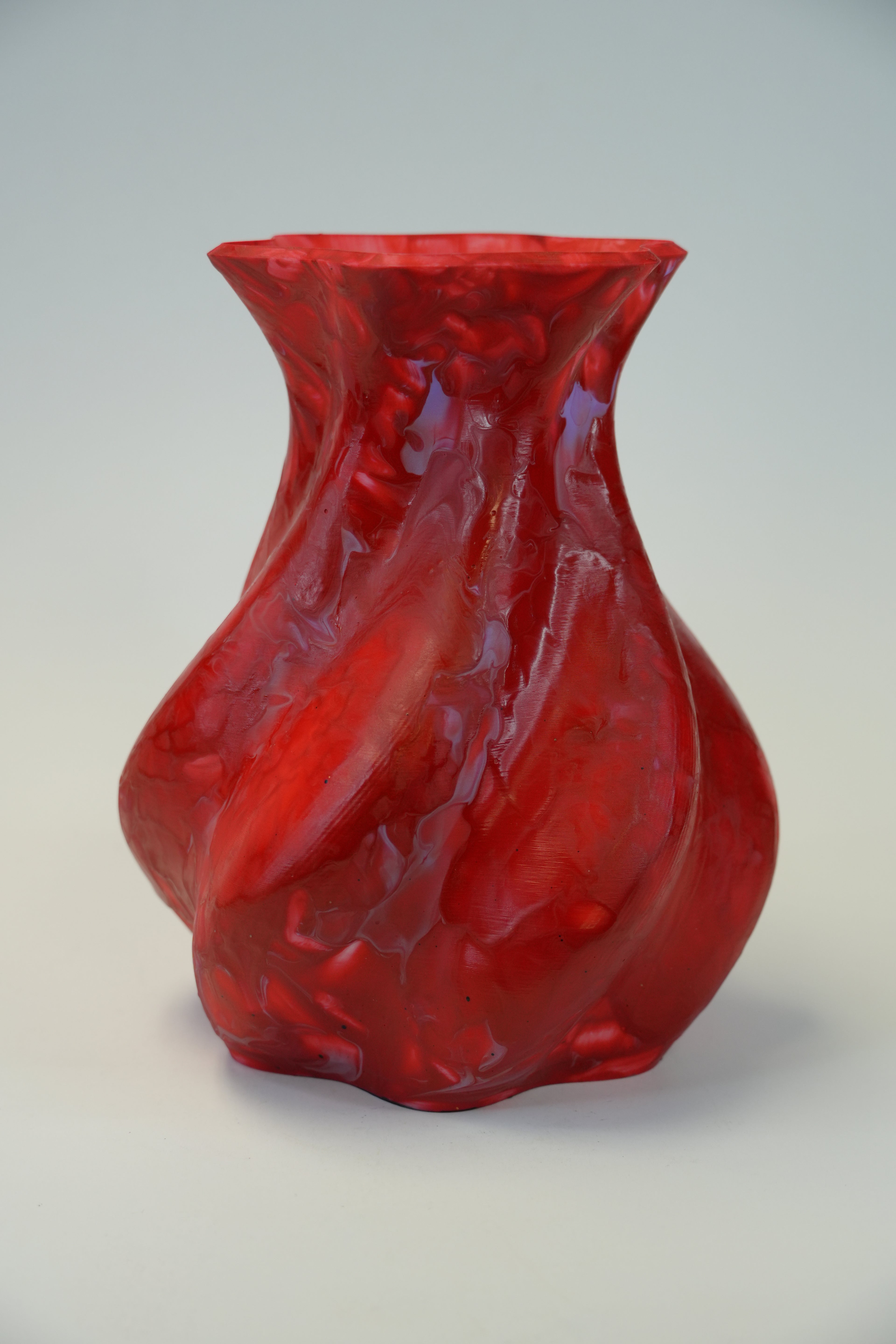 A translucent red vase with a sculptural, twisted organic shape and a flared rim, photographed against a neutral light gray background.