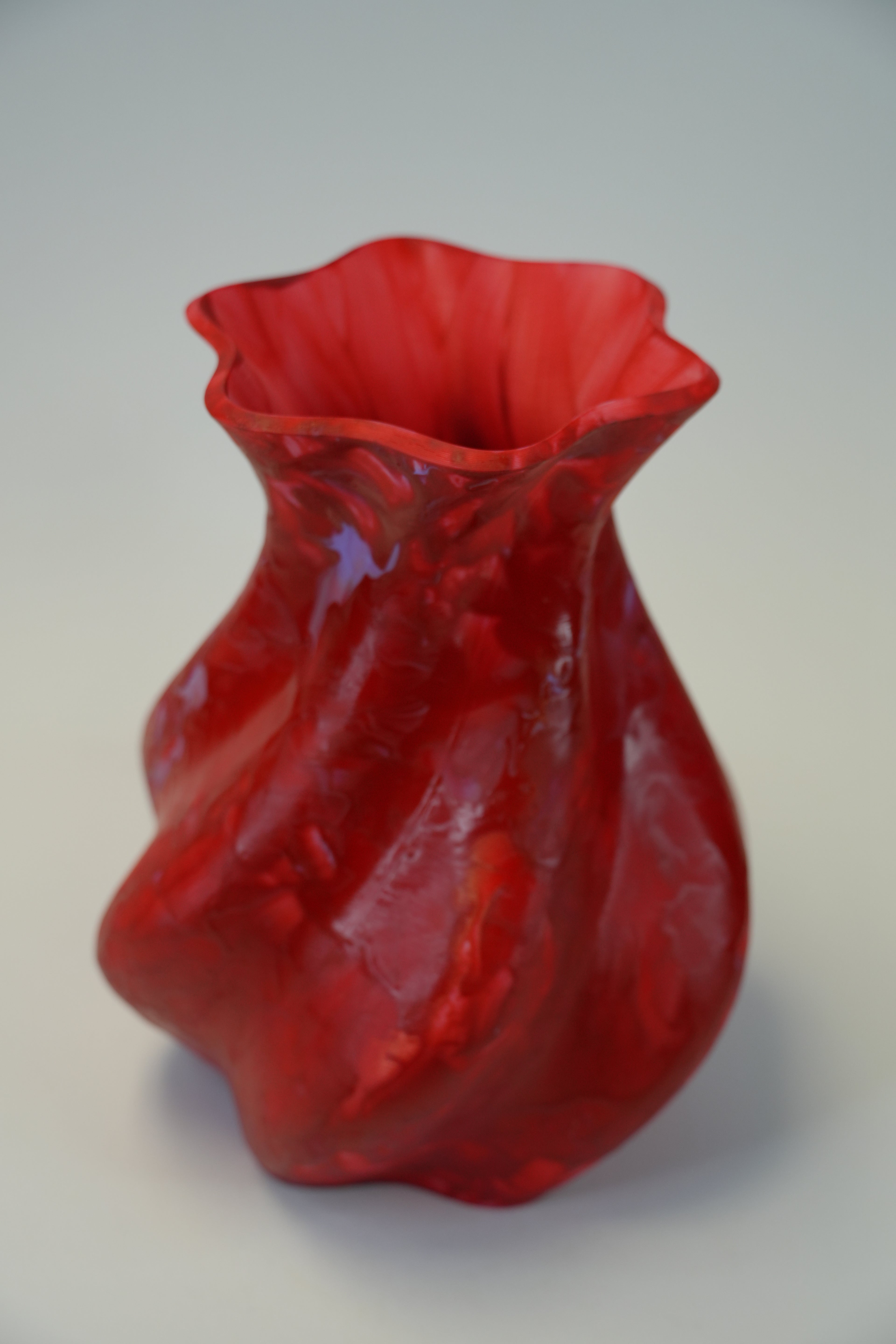 A sculptural, hand-blown glass vase in a vibrant translucent red, featuring a wavy, organic silhouette and a flared rim, set against a neutral light gray background.