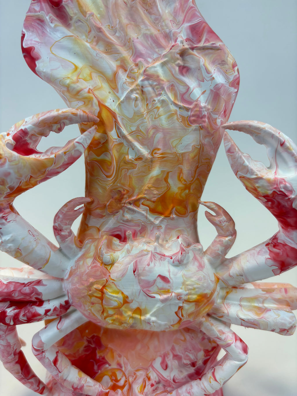 A close-up of a sculptural vase or decorative vessel shaped like a crab, featuring a marbled swirl pattern in white, pink, red, and amber-orange tones against a light gray background.