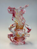 Marbled sculpture of a crab supporting a shell-like form, featuring swirls of pink, white, and orange-yellow on a neutral studio background.