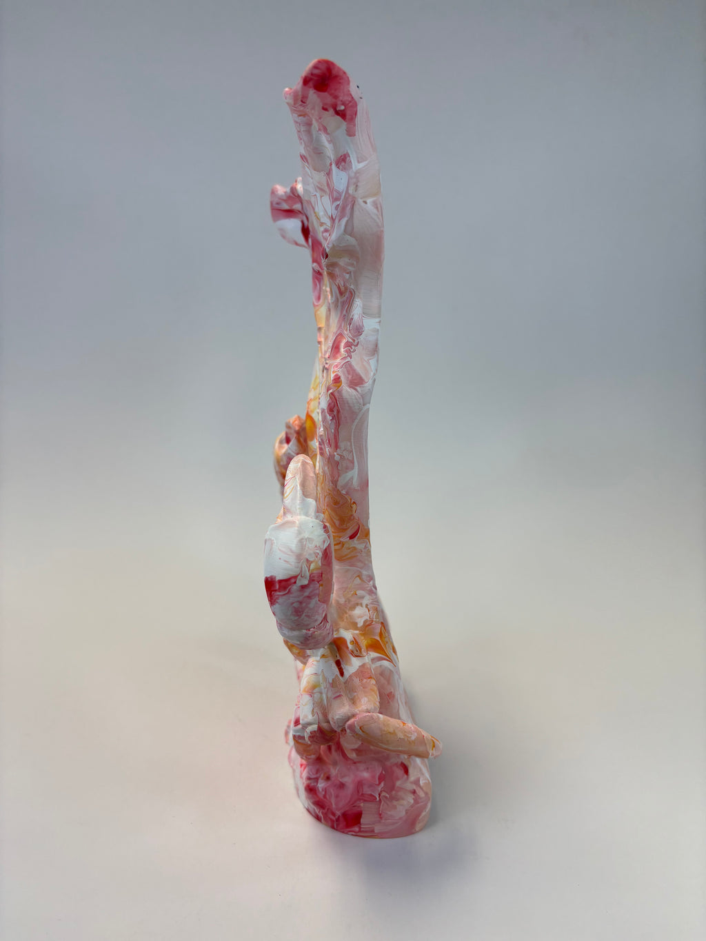 A tall, abstract sculptural decorative object with a marbled pattern of white, pink, red, and orange, photographed against a neutral light gray studio background.