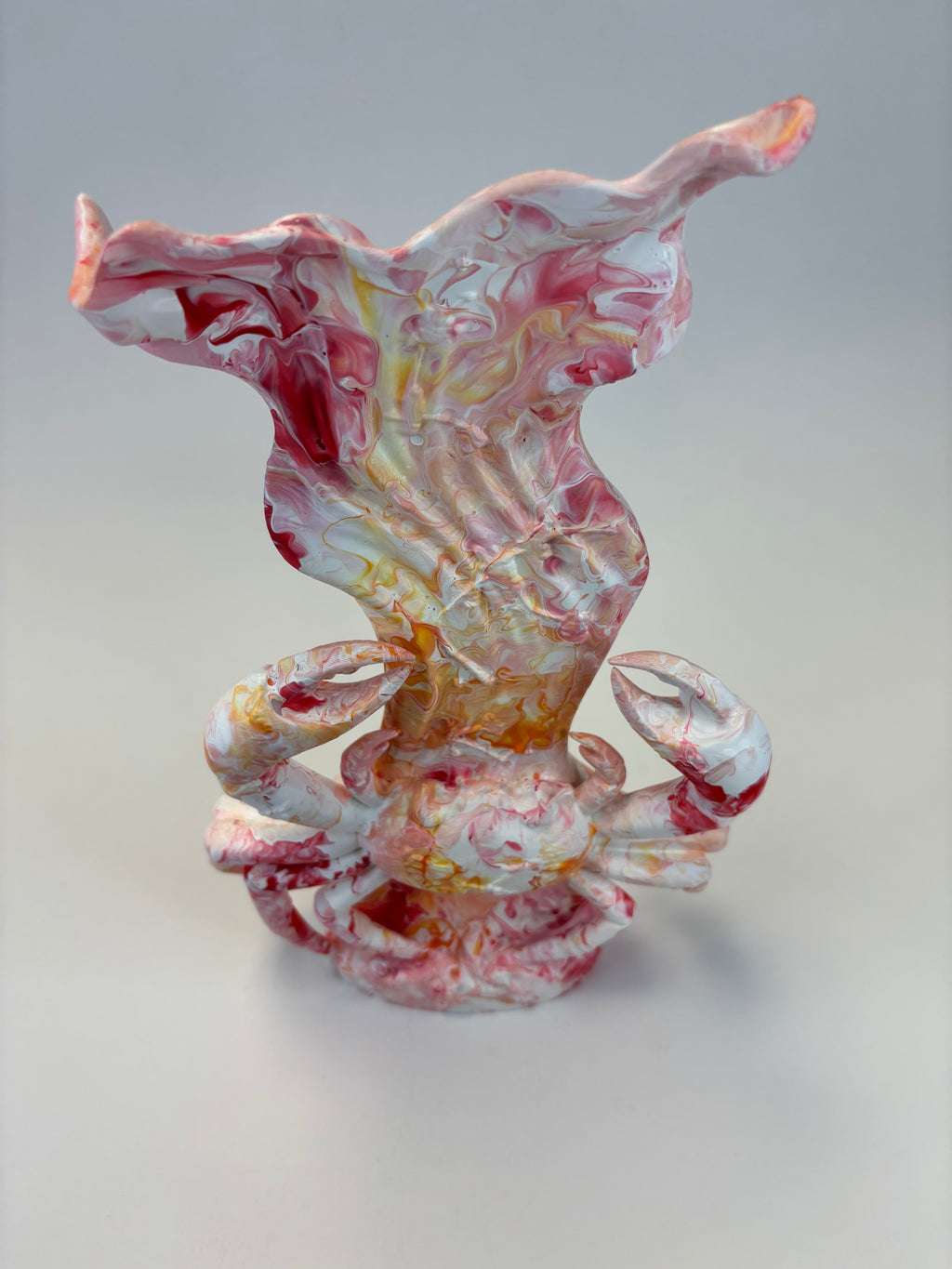 A sculptural piece featuring a stylized crab at the base with a flowing, shell-like form rising above it, in a marbled glaze of white, pink, red, and yellow on a light gray background.