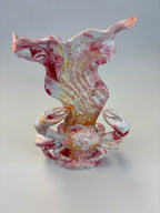 A sculptural piece featuring a stylized crab at the base with a flowing, shell-like form rising above it, in a marbled glaze of white, pink, red, and yellow on a light gray background.