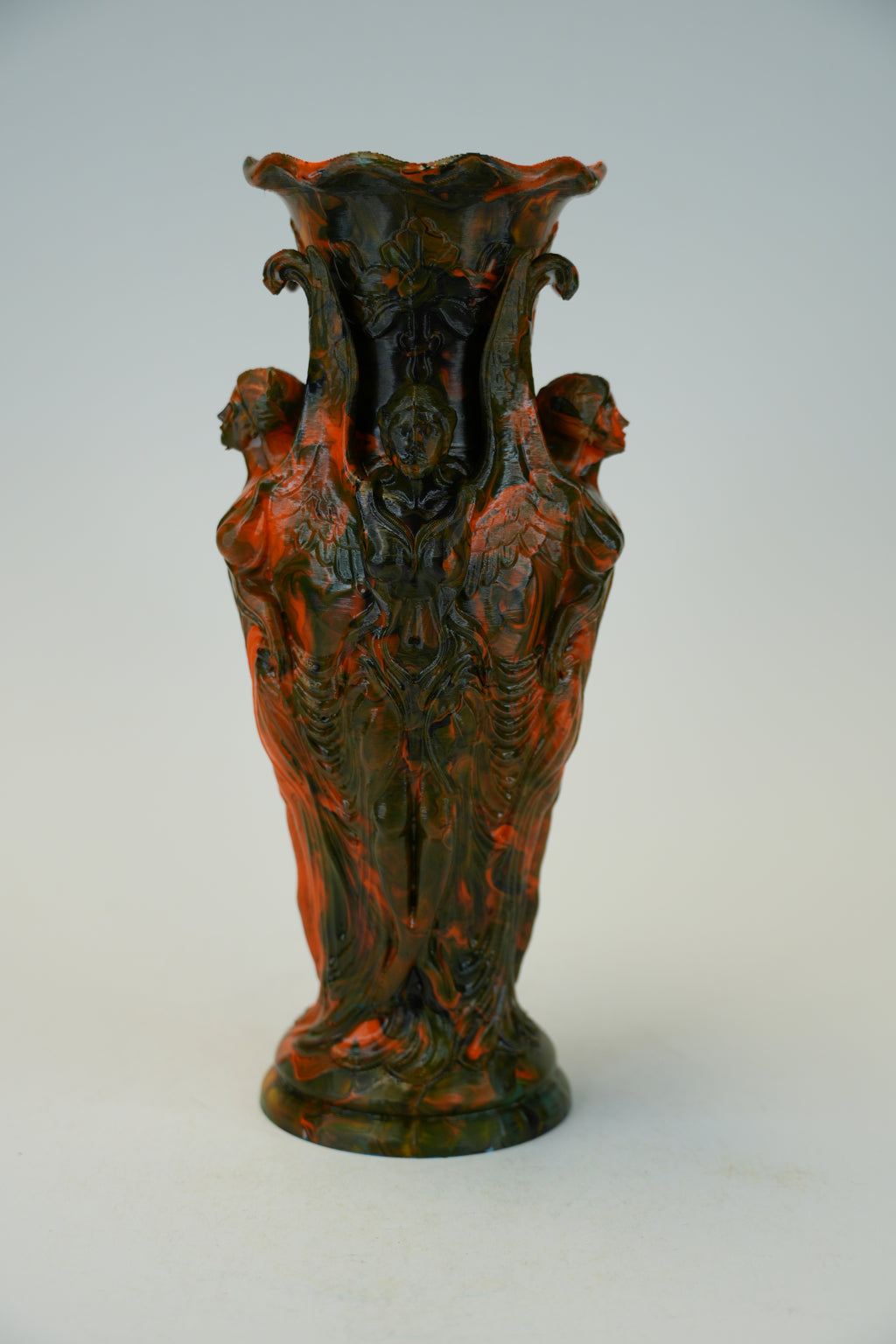 An ornate sculptural ceramic vase with a flared rim and four draped female figures, featuring a mottled dark green and terracotta red glaze, set against a plain light gray background.