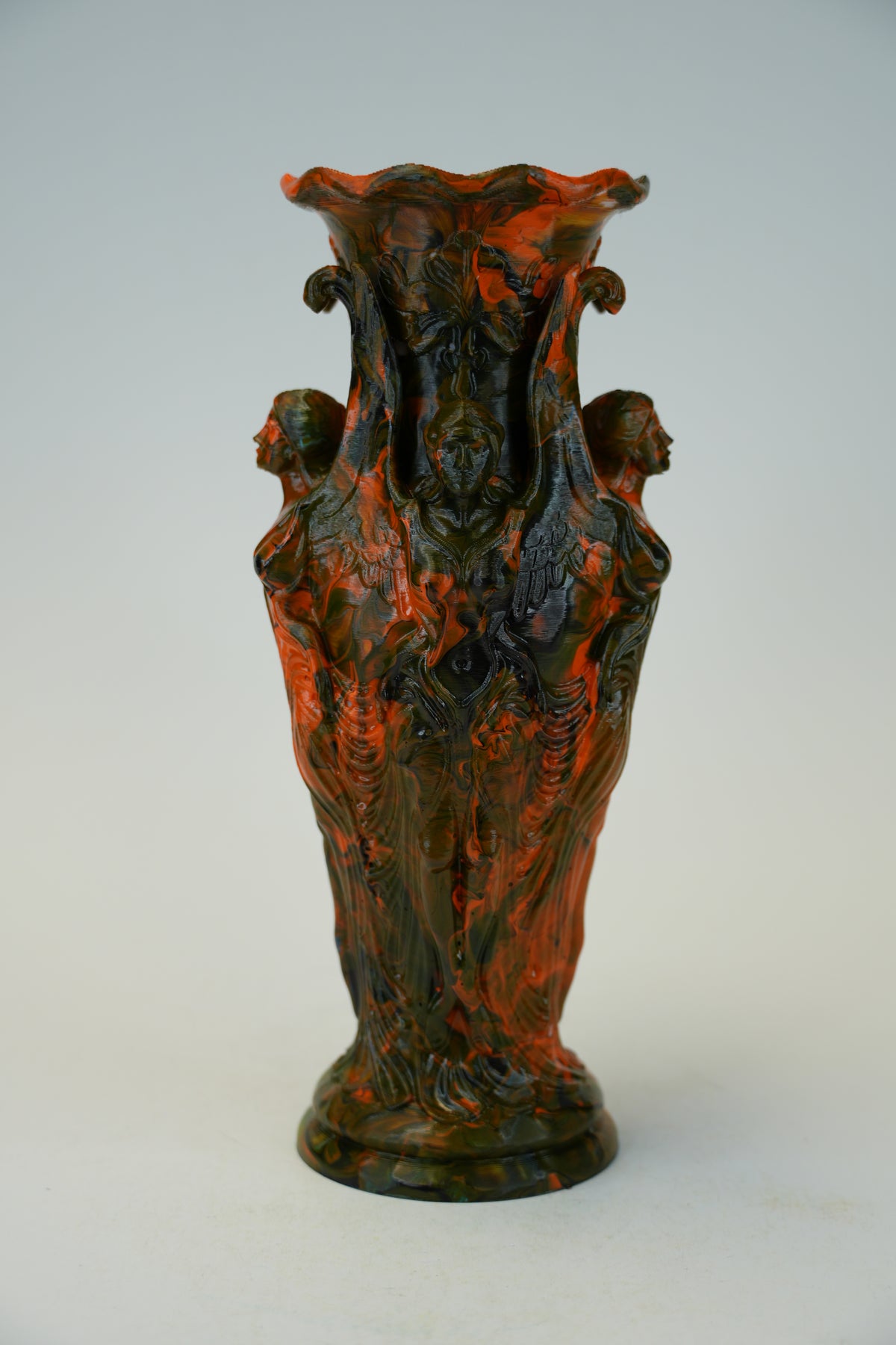 An ornate sculptural ceramic vase featuring four female figures with draped hair, rendered in a marbled mix of glossy orange-red and dark green glaze, set against a plain light gray background.