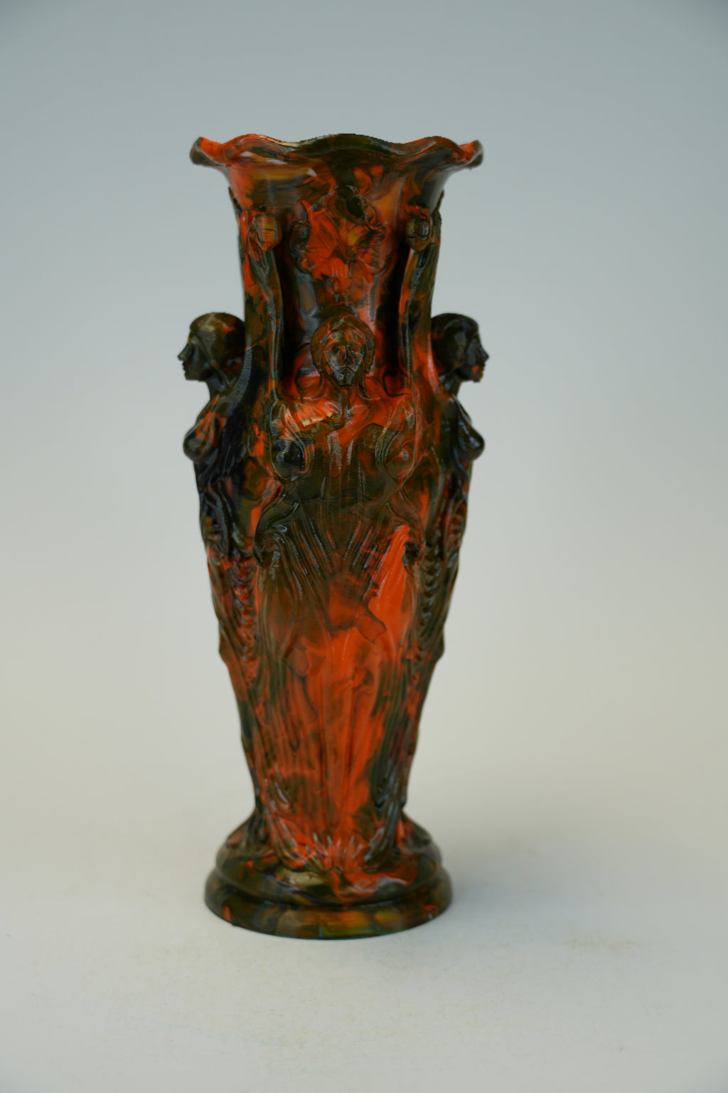An ornate sculptural ceramic vase with a flared rim and four draped female figures, featuring a mottled dark green and terracotta red glaze, set against a plain light gray background.
