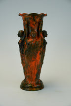 An ornate sculptural ceramic vase with a flared rim and four draped female figures, featuring a mottled dark green and terracotta red glaze, set against a plain light gray background.
