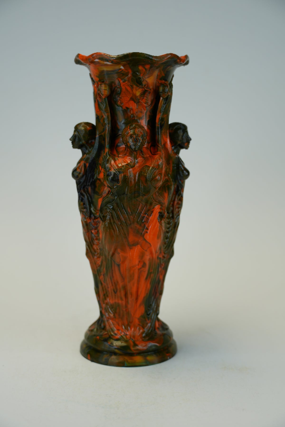 An ornate sculptural ceramic vase with a flared rim and four draped female figures, featuring a mottled dark green and terracotta red glaze, set against a plain light gray background.