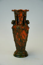 An ornate sculptural ceramic vase with a flared rim, featuring relief carvings of draped figures and two side faces, finished in a mottled dark green and burnt orange glaze.