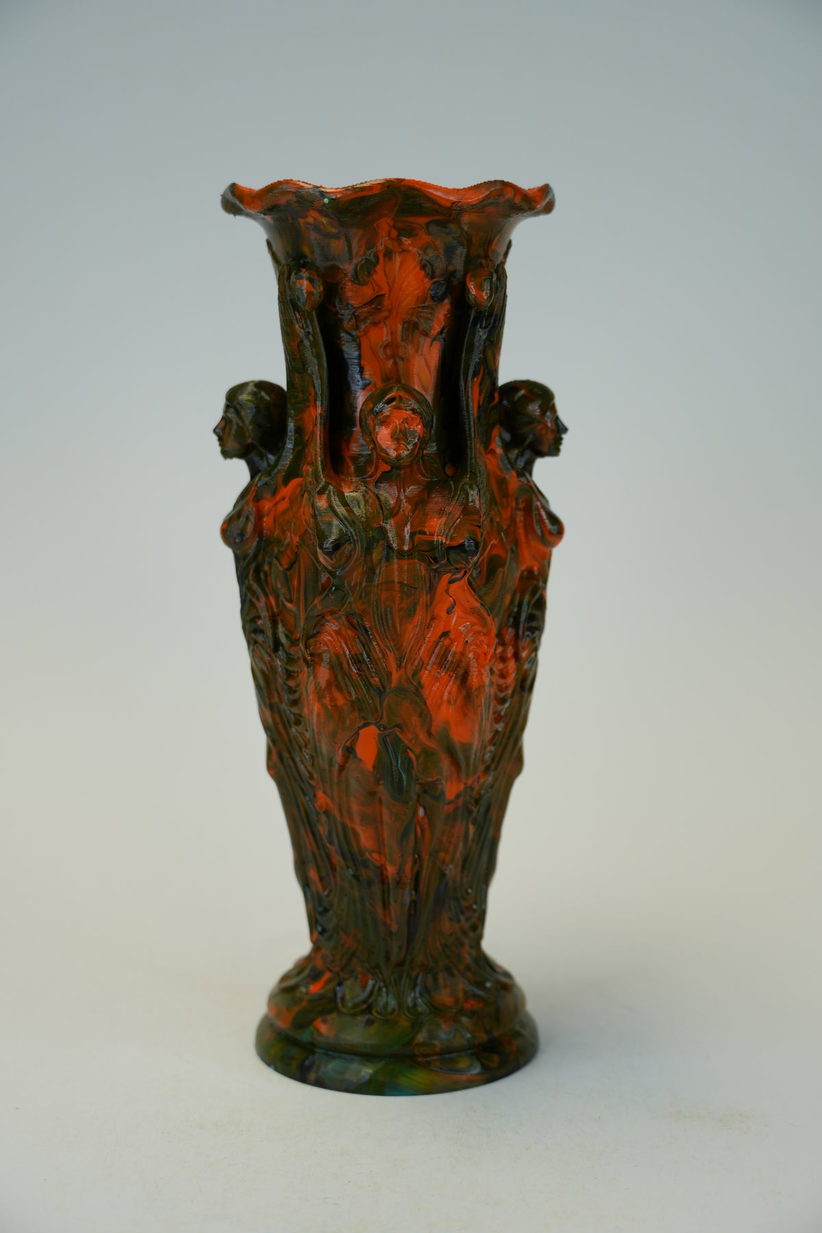 An ornate sculptural ceramic vase with a flared rim, featuring relief carvings of draped figures and two side faces, finished in a mottled dark green and burnt orange glaze.