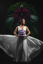 Woman in a white dress with a purple floral detail against a dark background with large leaves.