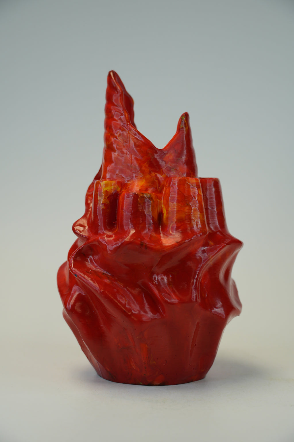 A glossy red sculptural vase with a textured, organic flame-like form and a flared rim, photographed against a neutral light gray studio background.