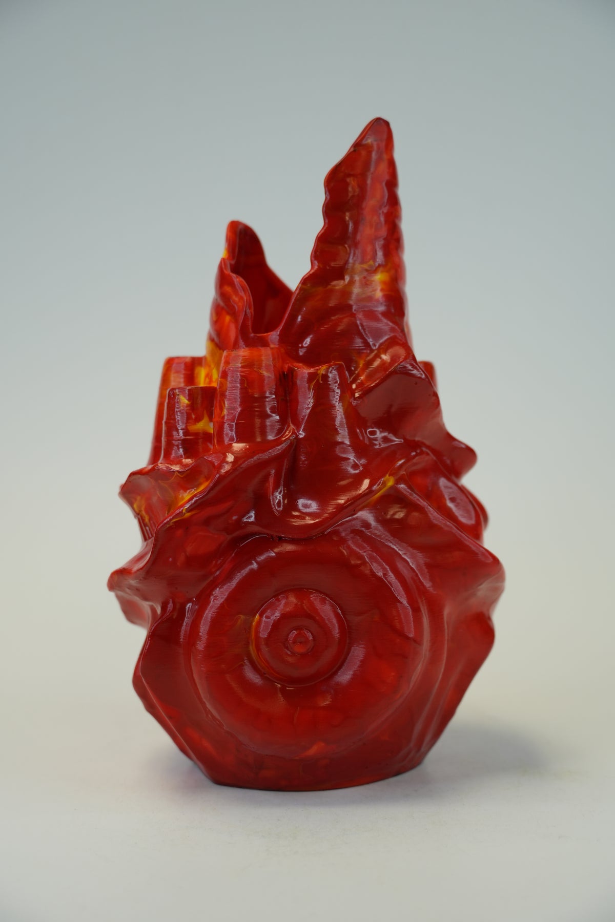 A glossy red translucent sculptural object with a spiral shell-like base and jagged pointed top, photographed against a plain light gray studio background.