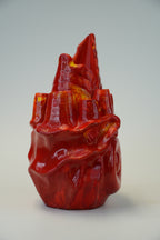 A glossy red sculptural object with a highly textured, organic, and flame-like form, photographed against a neutral light gray background.