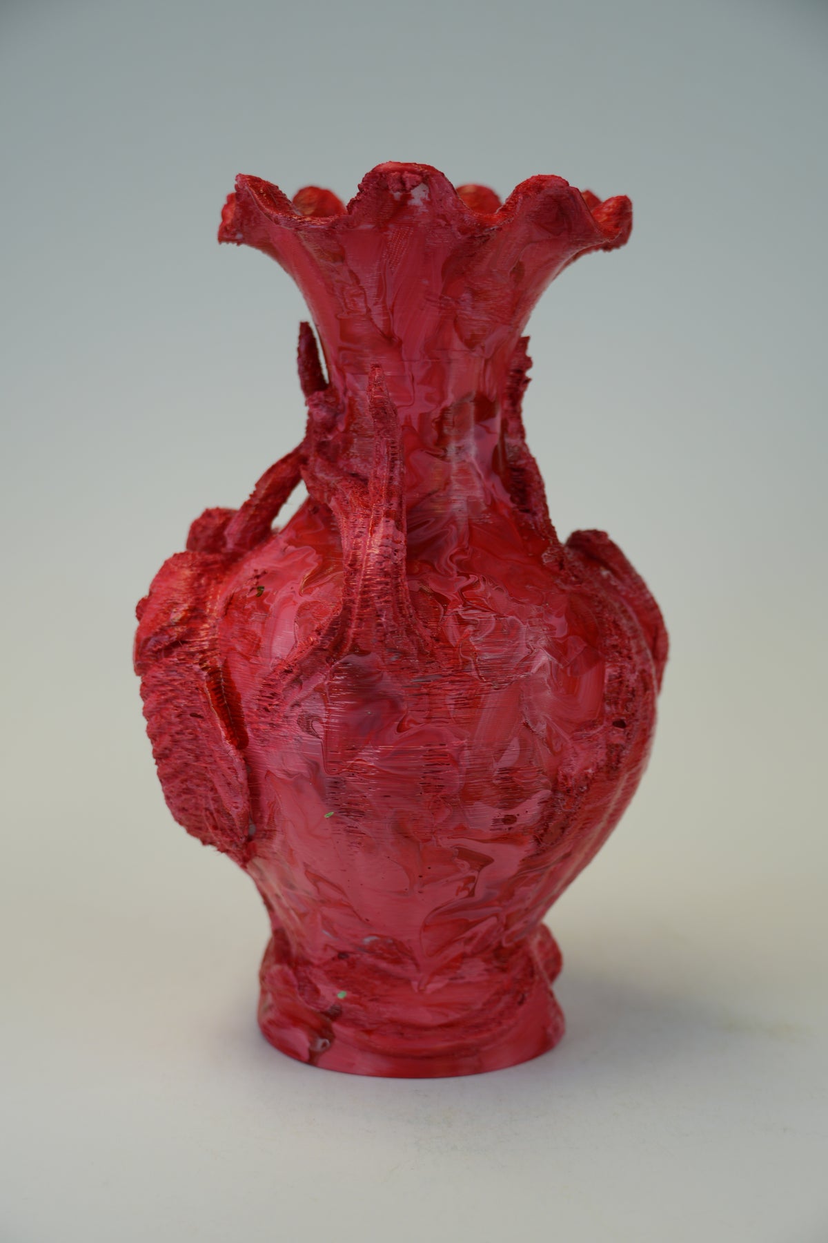 A textured, sculptural red ceramic vase with a flared, ruffled neck and a bulbous body, photographed against a neutral light gray background.