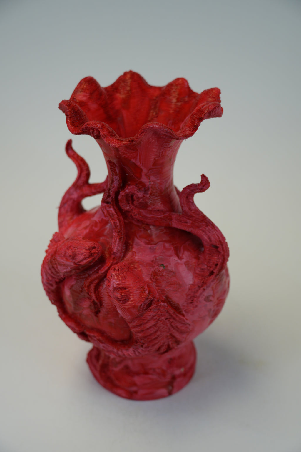 A red sculptural ceramic vase with a flared ruffled rim, featuring raised dragon-like figures wrapping around the body, photographed on a plain light gray background.
