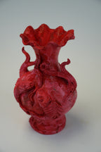 A red sculptural ceramic vase with a flared ruffled rim, featuring raised dragon-like figures wrapping around the body, photographed on a plain light gray background.