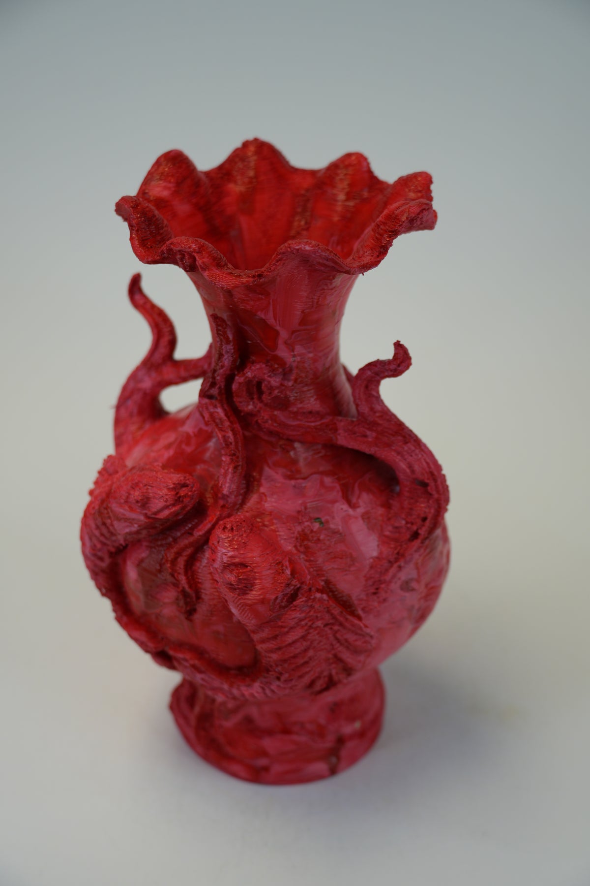 A red sculptural ceramic vase with a flared ruffled rim, featuring raised dragon-like figures wrapping around the body, photographed on a plain light gray background.