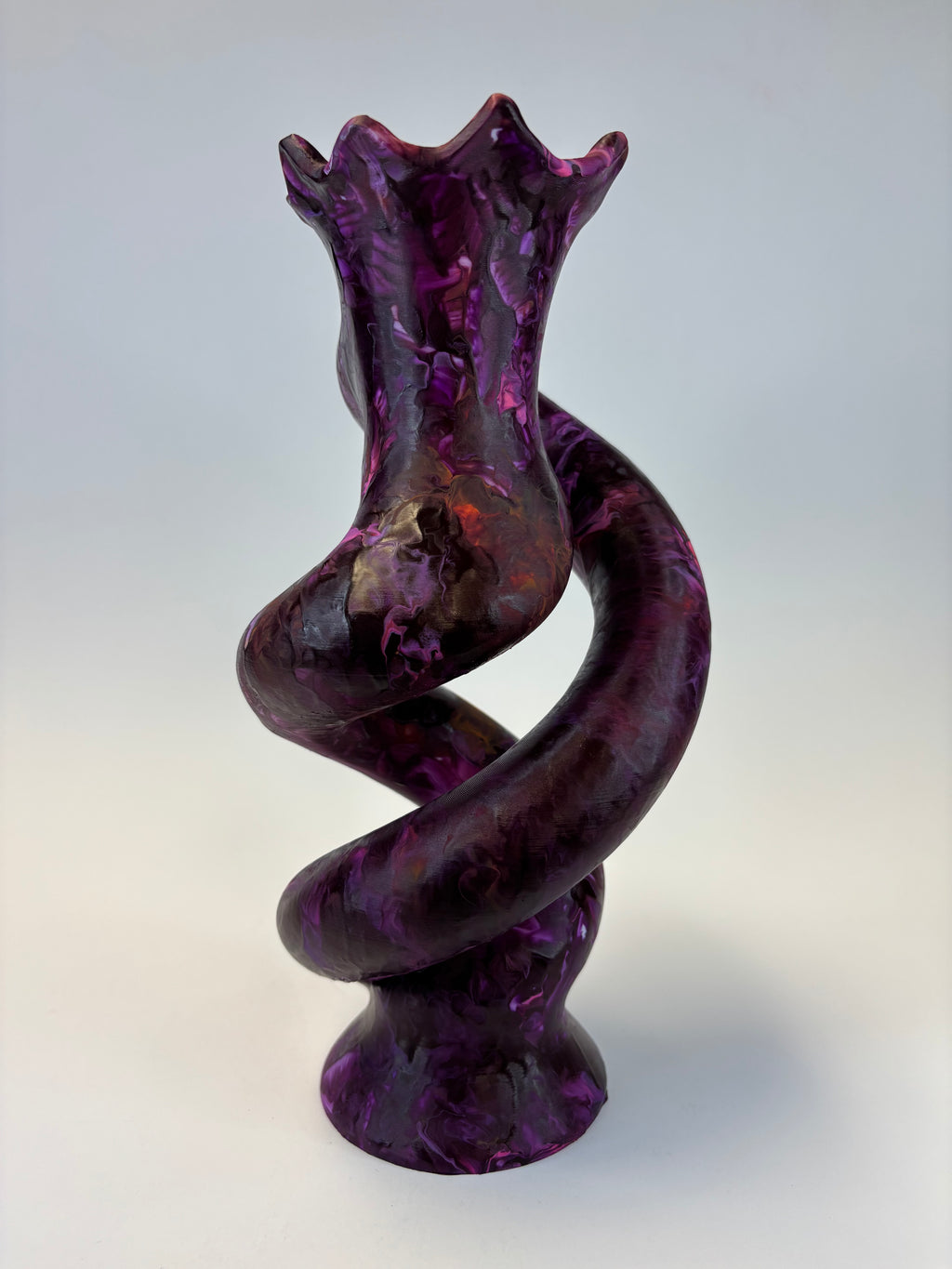 A sculptural spiral object made of glossy, marbled purple glass with darker shading, set against a neutral light gray background.
