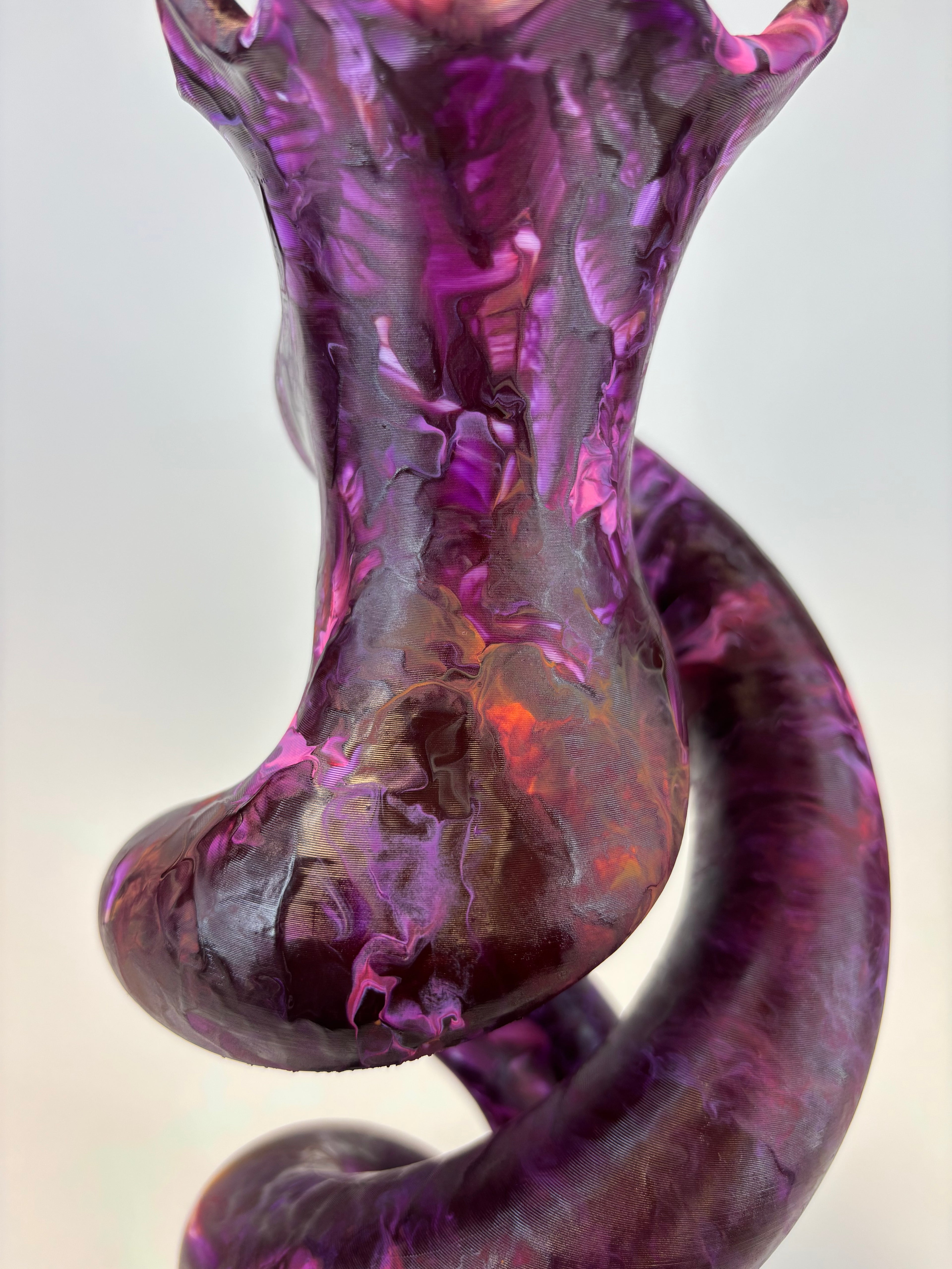 A close-up of a glossy abstract sculptural object with swirling purple, magenta, and dark marbled patterns, photographed against a soft light gray background.