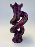 A sculptural twisted object with a marbled purple, pink, and dark brown pattern, photographed against a light gray background.