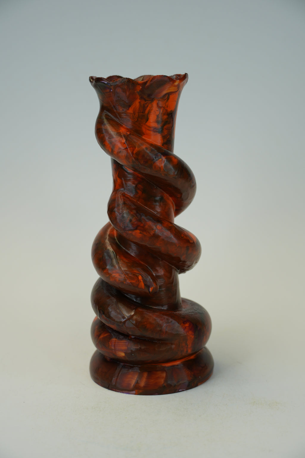 A sculptural spiral glass vase made of amber-brown marbled glass, standing on a round base against a light gray studio background.