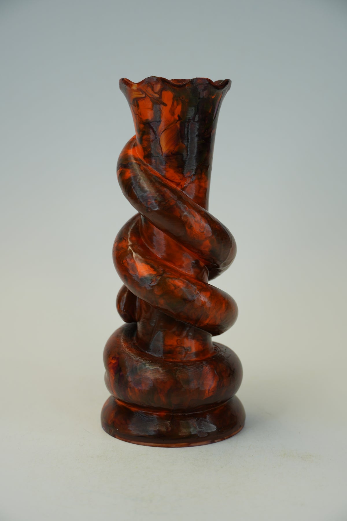 A sculptural spiral vase made of glossy amber-brown glass with darker marbled streaks, photographed against a plain light gray studio background.