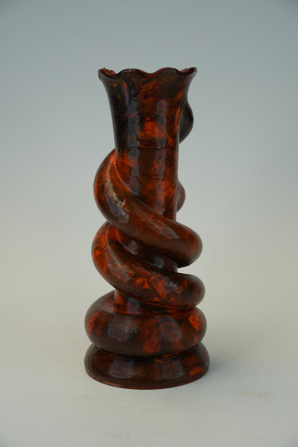 A sculptural spiral vase made of glossy amber-brown glass with darker marbling, photographed against a plain light gray studio background.