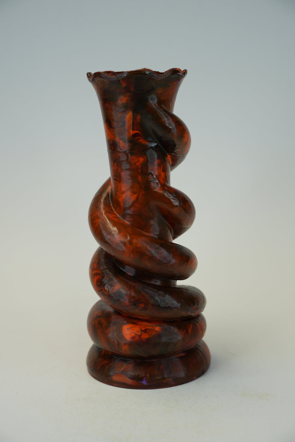 A sculptural spiral glass vase with a flared rim, featuring a glossy amber-brown and black marbled pattern, set against a neutral light gray background.