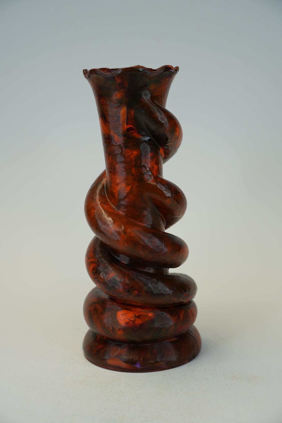 A sculptural spiral glass vase with a flared rim, featuring a glossy amber-brown and black marbled pattern, set against a neutral light gray background.