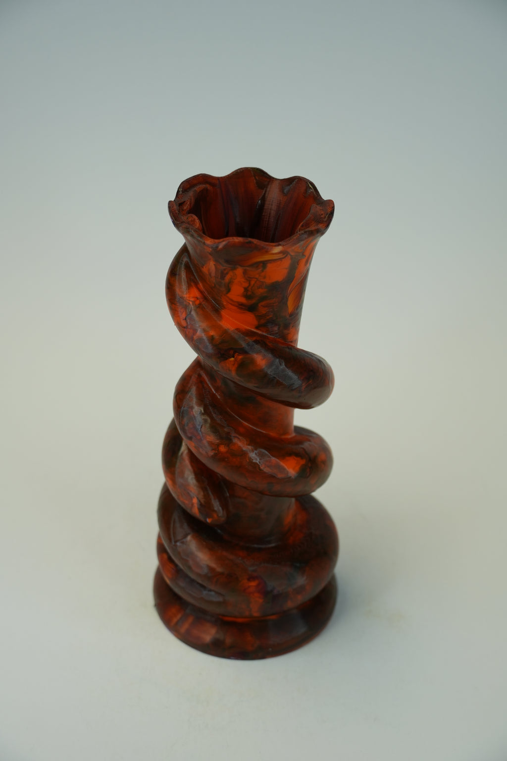 A small sculptural vase made of glossy amber-brown translucent glass with a spiral twisted form and a flared rim, photographed against a plain light gray studio background.
