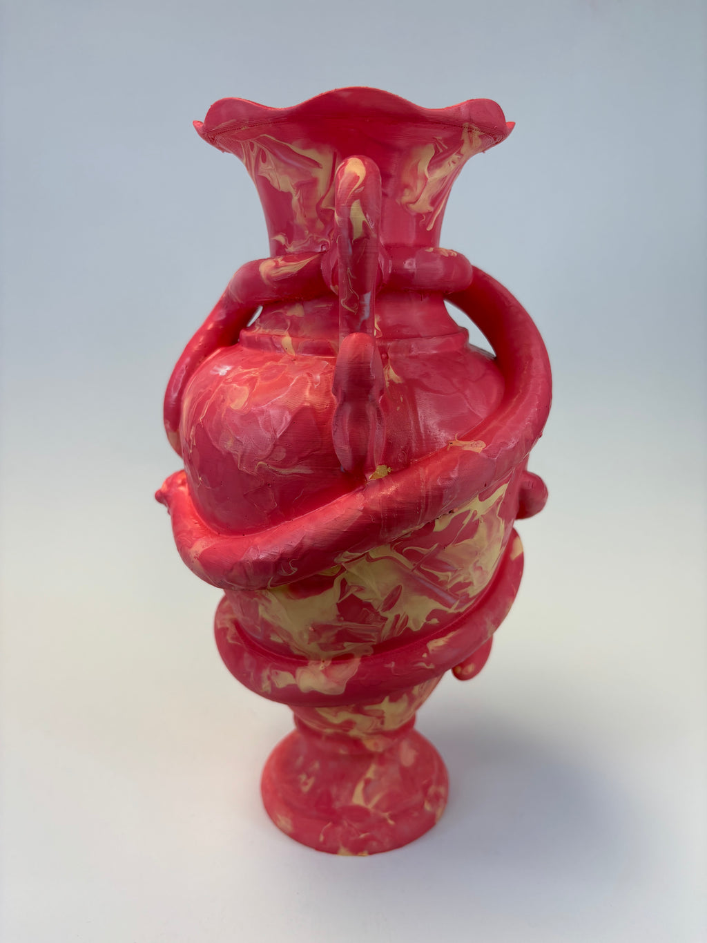 A sculptural vase with a bright pink and beige marbled pattern, featuring a flared rim, looped handles, and a pedestal base, photographed against a light gray studio background.