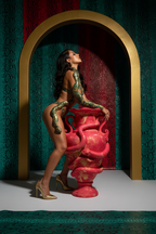 A model wearing a green snakeskin-patterned lingerie set poses next to a large pink decorative urn in front of an arched gold frame against a dark green patterned wall.