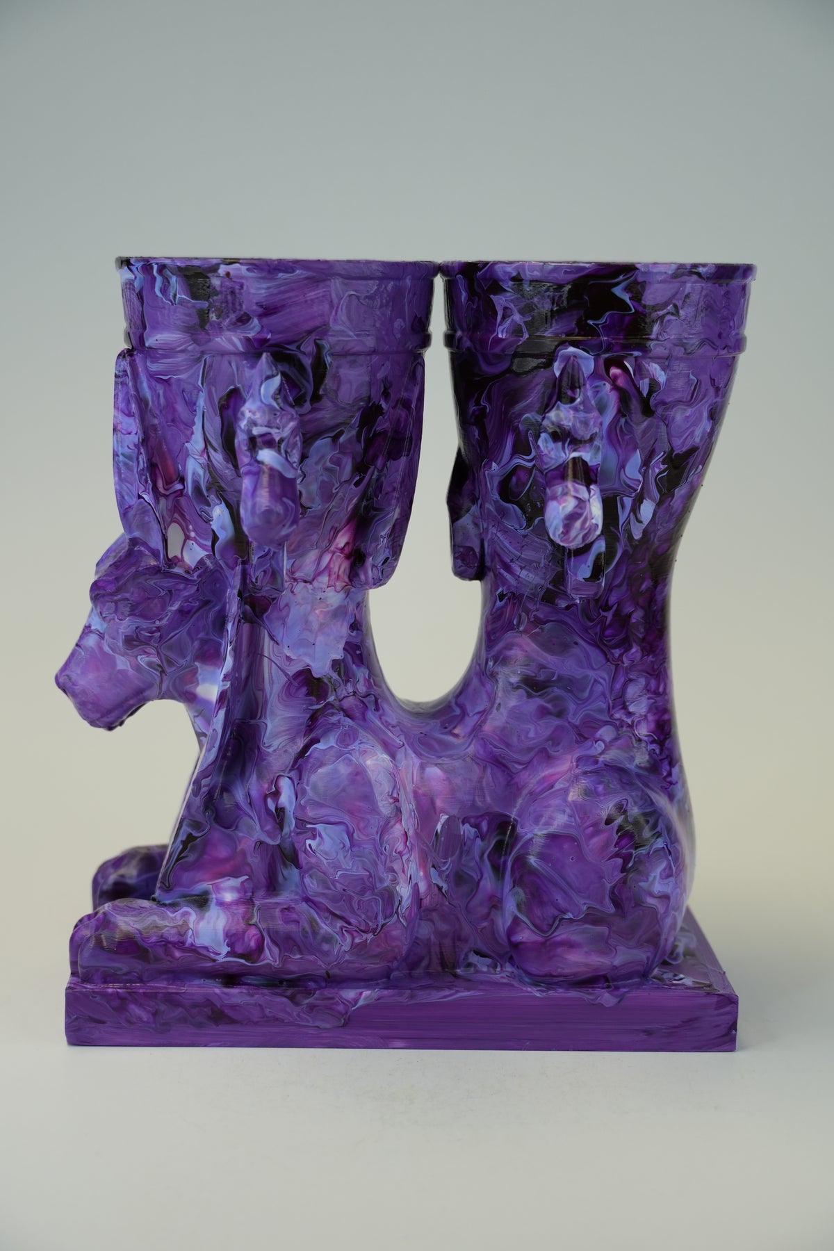 A sculptural purple and black marbled vase or candle holder with a hollow center and a rectangular base, photographed against a plain light gray studio background.