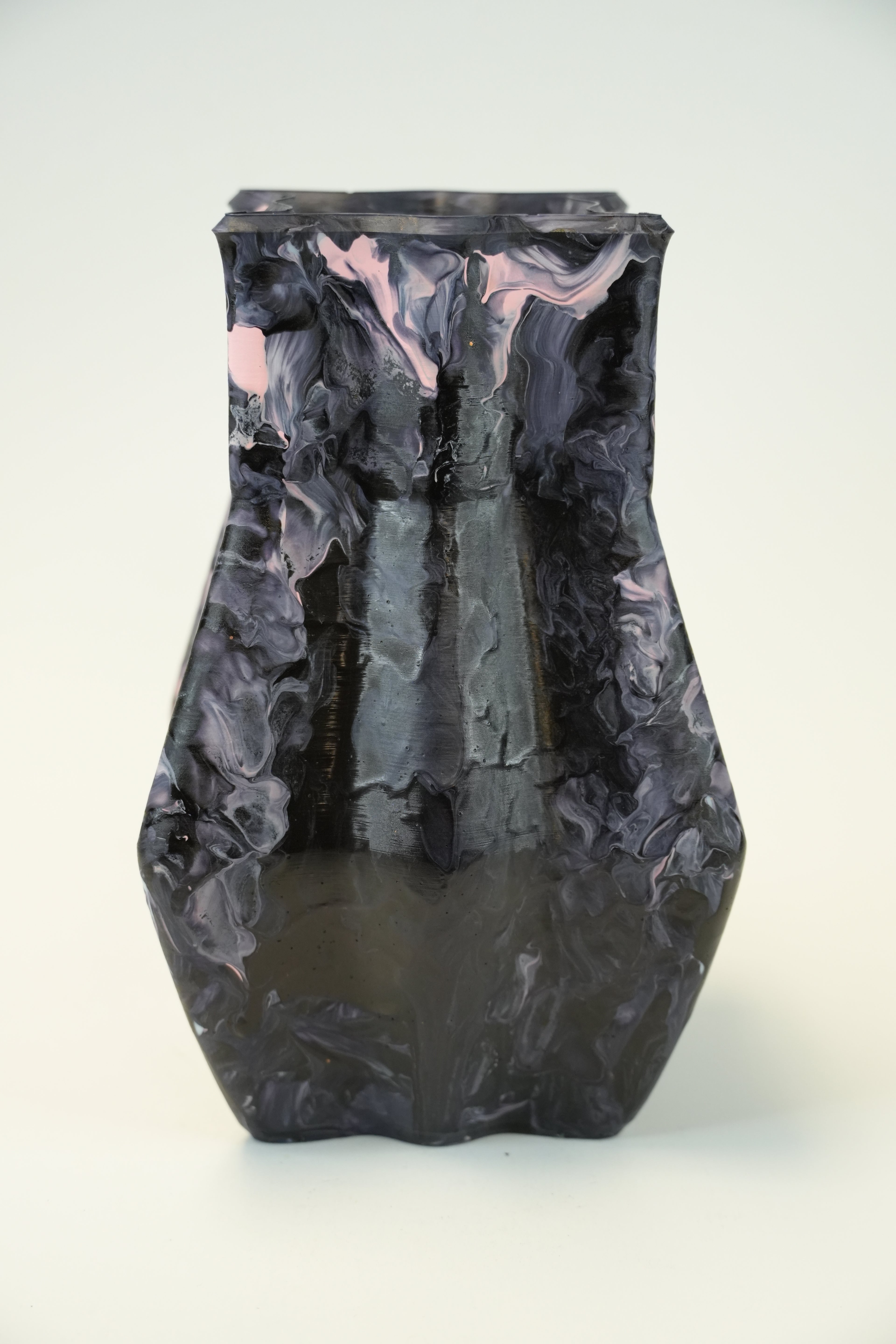 A dark, abstract sculptural vase with a glossy, marbled finish in black, charcoal gray, and subtle pink highlights, set against a plain off-white background.