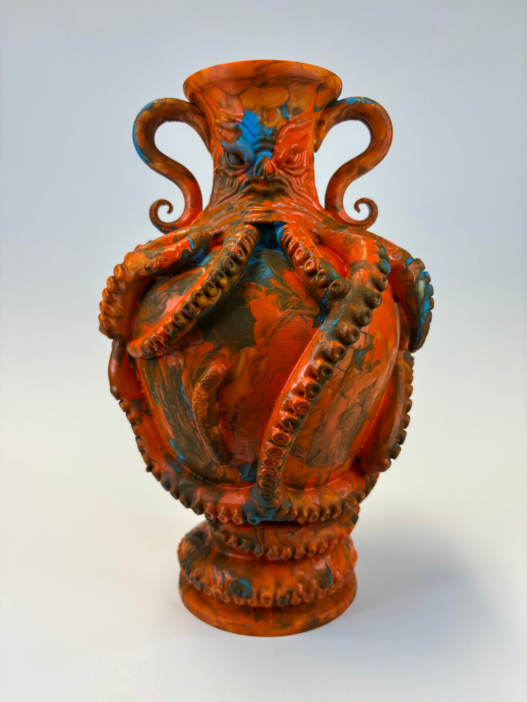 An ornate sculptural vase with a glossy terracotta orange glaze, featuring octopus tentacles wrapping around the body and handles, accented with blue and dark brown details, set against a light gray background.