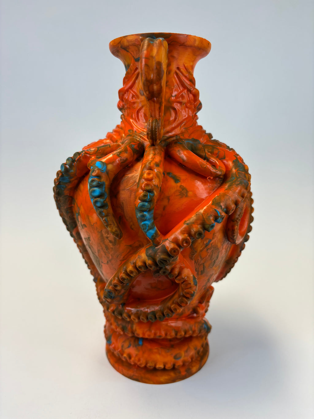 An orange vase with a textured, organic surface resembling a stylized octopus or tentacle design, featuring blue accents and dark brown details, set against a light gray background.